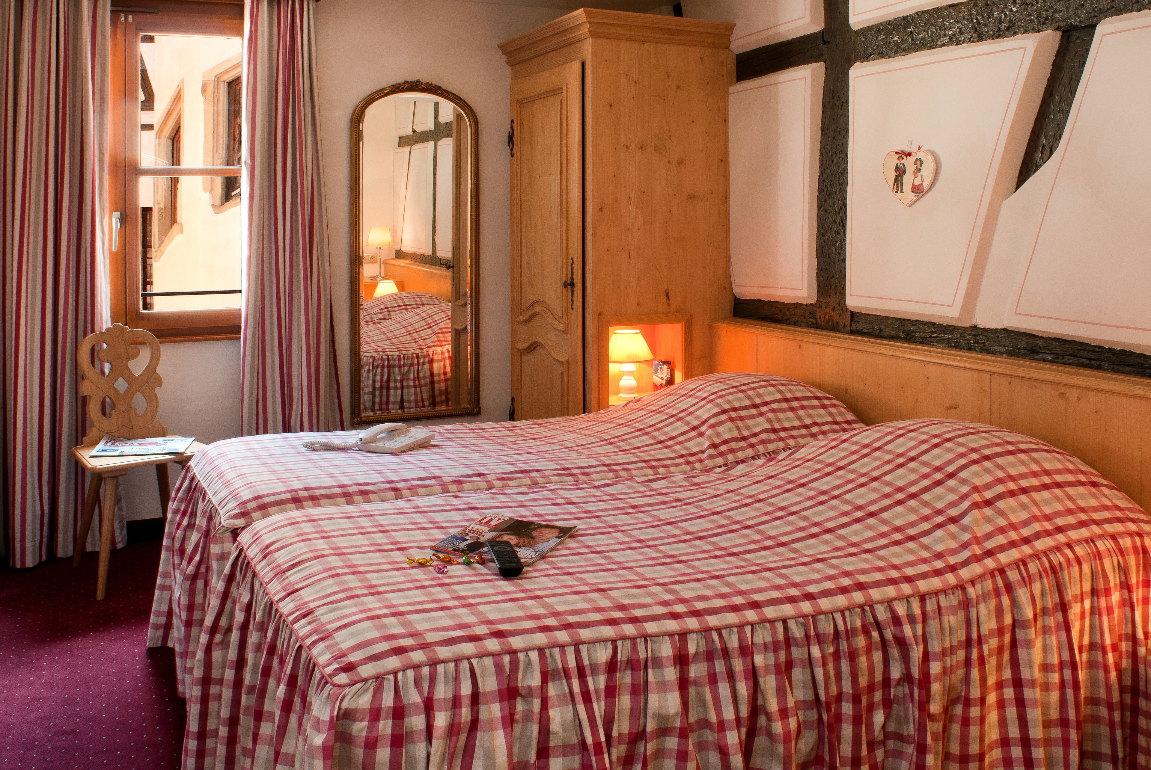 Hotellihuone, jossa on ruutukuviollinen peitto, puinen vaatekaappi, peili ja ikkuna, jossa on verhot.