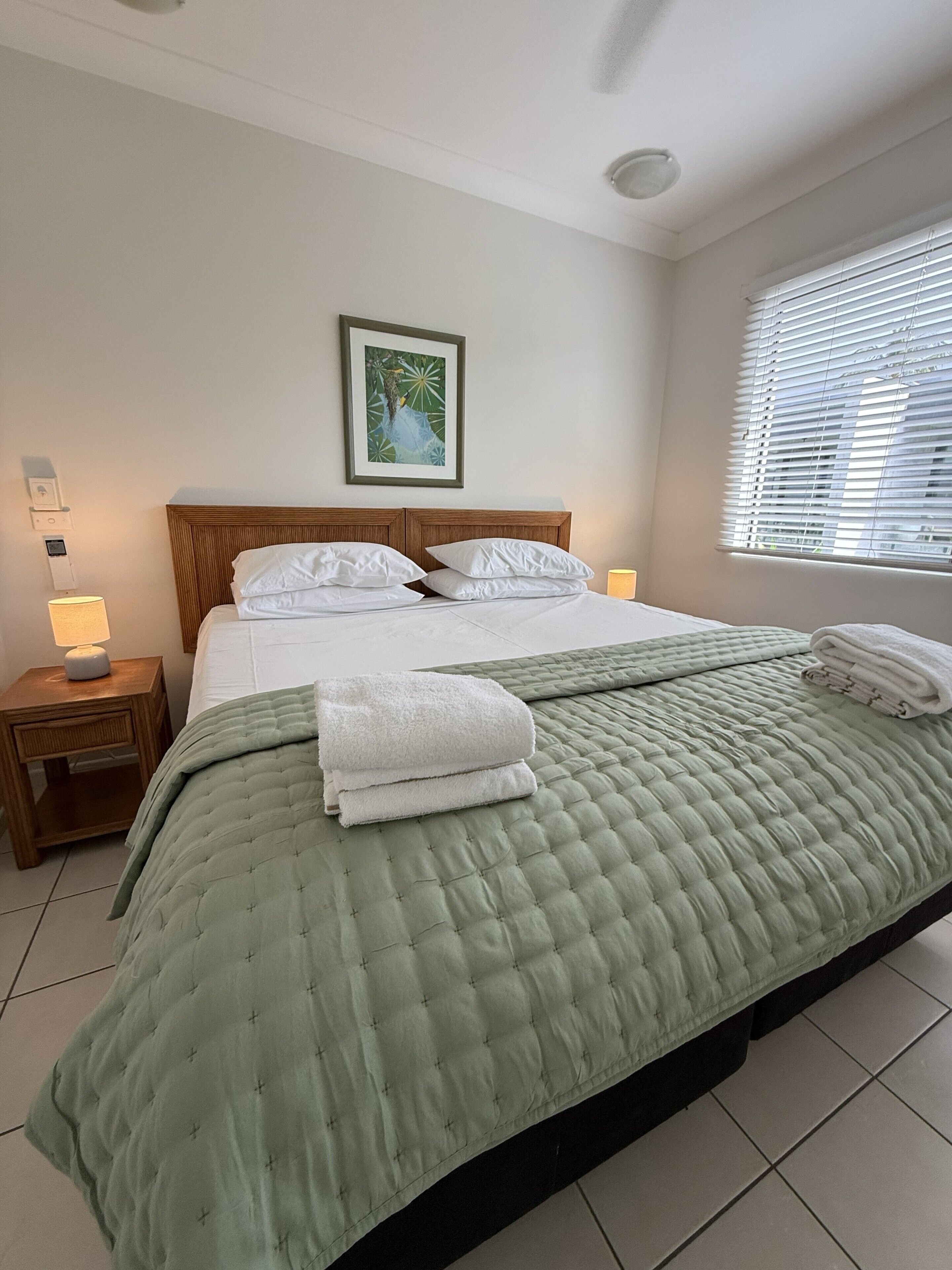 A neatly made bed with a green quilt, white pillows, and folded towels on a wooden headboard.