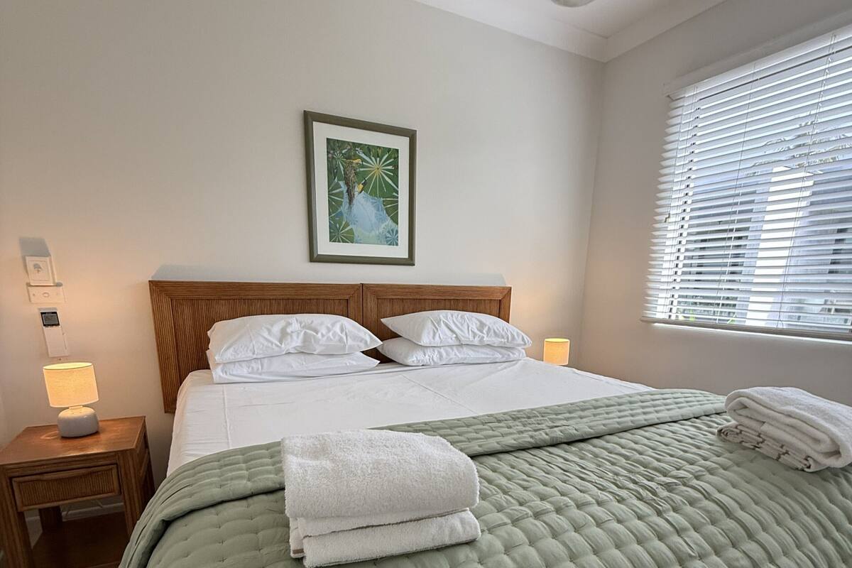 A neatly made bed with a green quilt, white pillows, and folded towels on a wooden headboard.