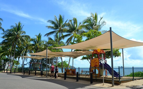 A playground with a slide and swings, shaded by large canopies, and surrounded by palm trees. - Melaleuca Resort (Palm Cove)