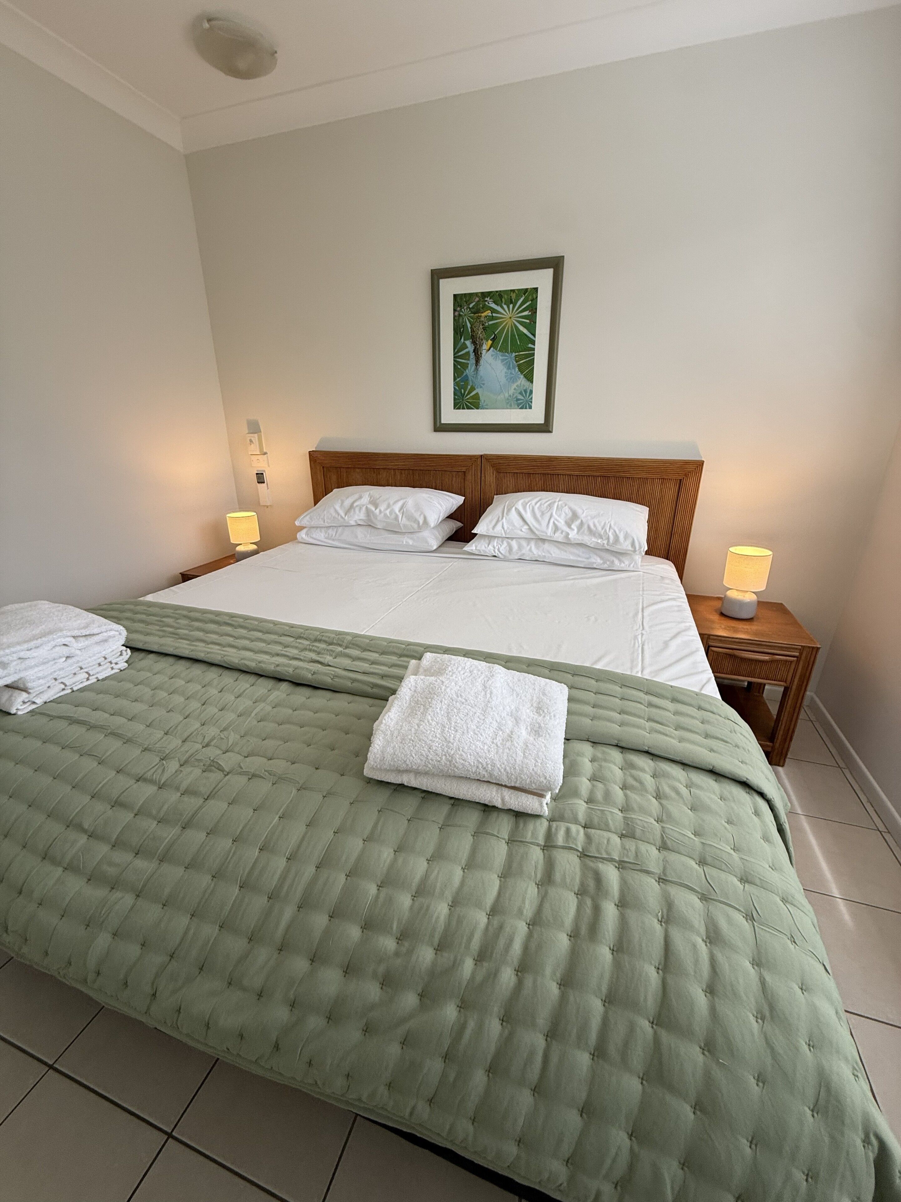 Un lit bien fait, avec une couette verte, des oreillers blancs et une serviette blanche pliée posée sur le lit. Un tableau encadré est accroché au mur, au-dessus du lit.