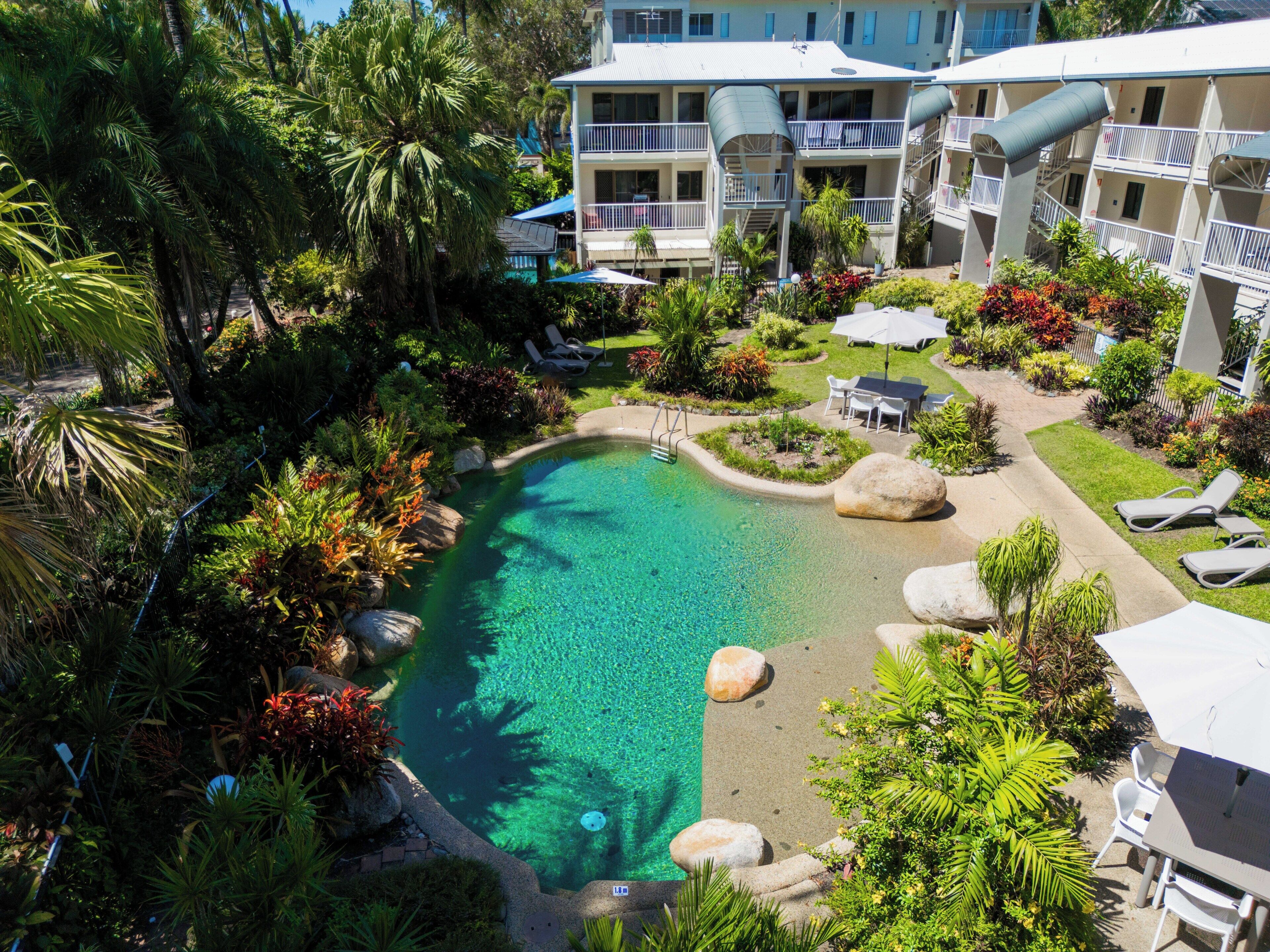 A resort with a swimming pool surrounded by tropical vegetation and a multi-story building with balconies.