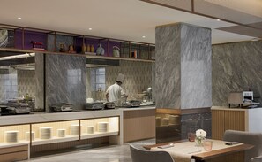 A modern restaurant with a chef preparing food, marble walls, and a buffet area with stacked plates. - Astoria Hotel (Mumbai)