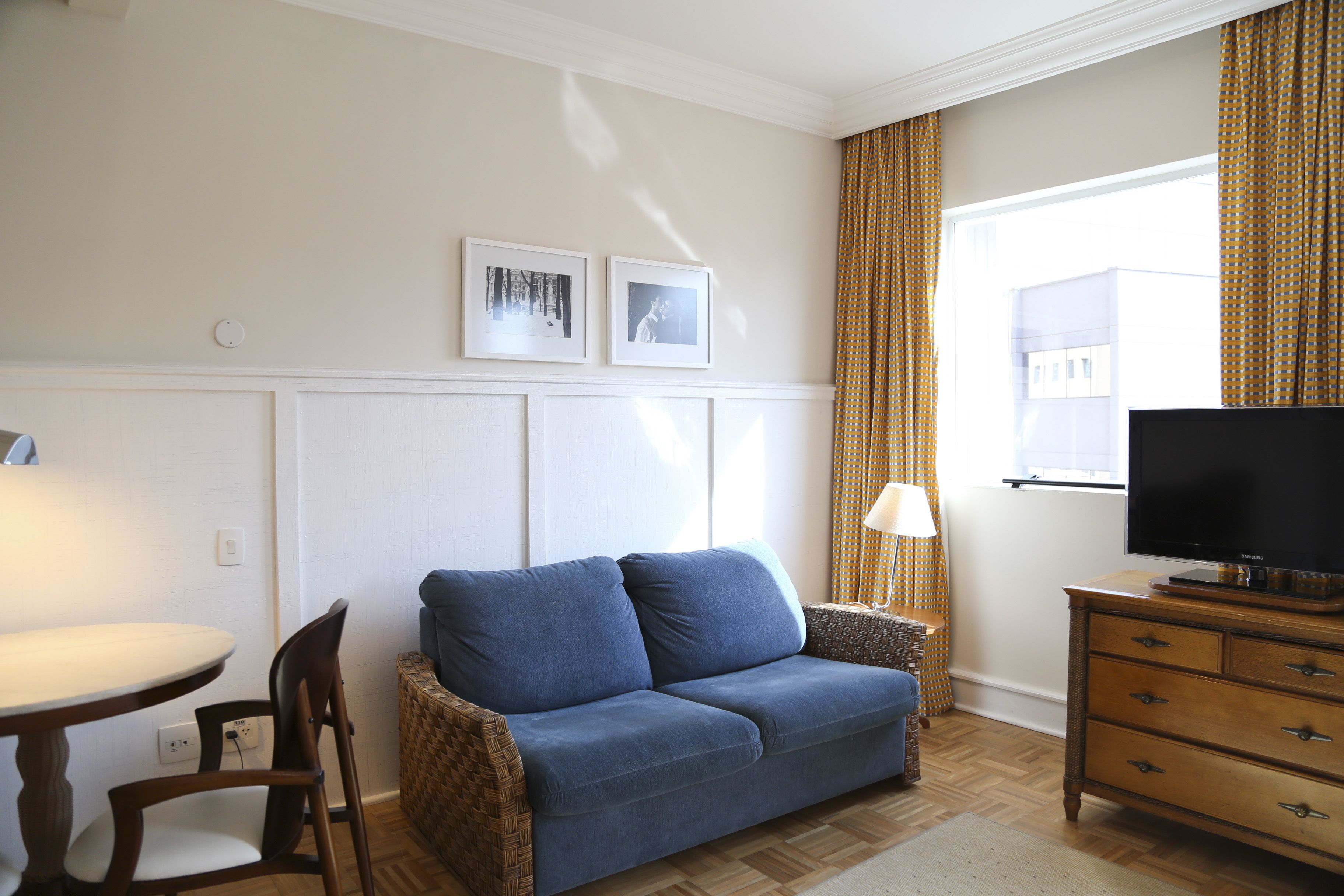 A living room with a blue sofa, a television on a wooden cabinet, a dining table with chairs, and two framed pictures on the wall.