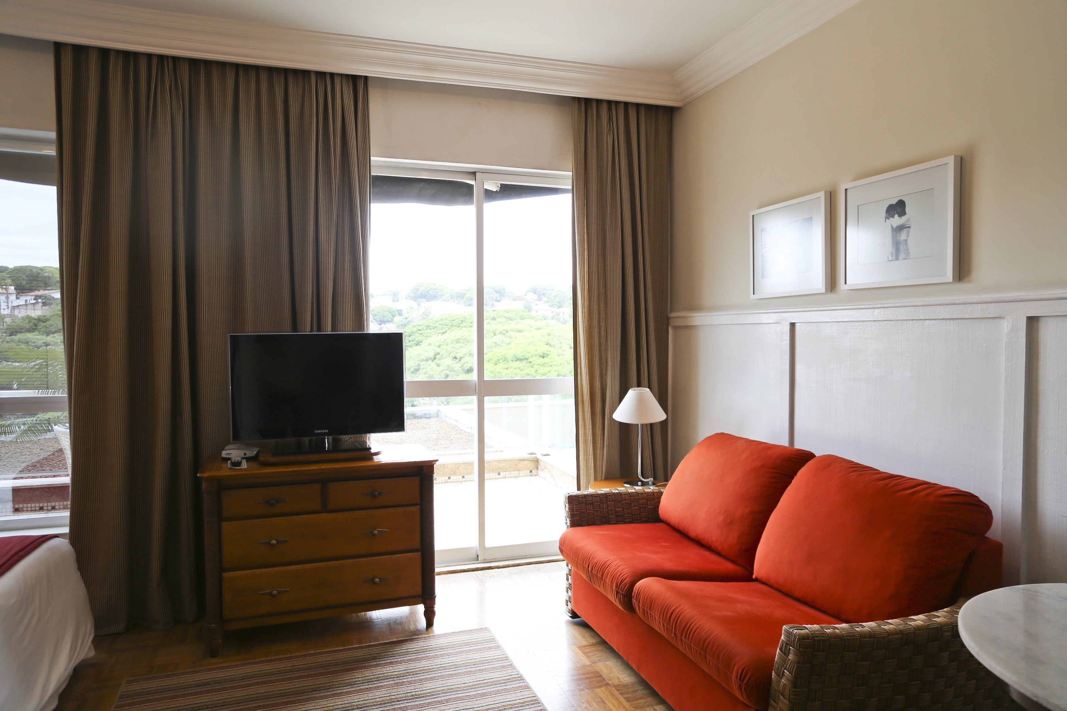 A hotel room with a television on a wooden dresser, a red sofa, a small table, and a view of greenery through the window.