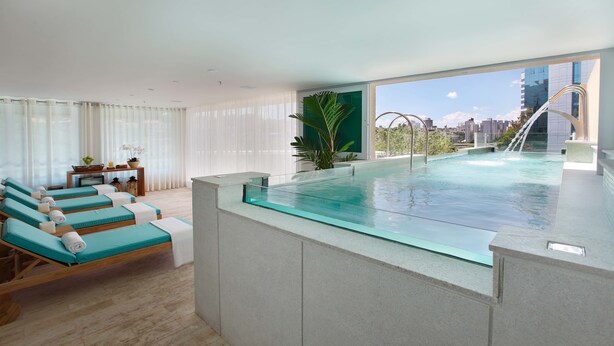 Une chambre d’hôtel moderne dotée d’une piscine à débordement avec des parois en verre, d’une terrasse ensoleillée avec des chaises longues et d’une vue sur la ville.
