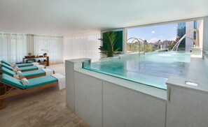 Une chambre d’hôtel moderne dotée d’une piscine à débordement avec des parois en verre, d’une terrasse ensoleillée avec des chaises longues et d’une vue sur la ville.