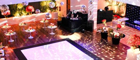 A spacious event hall with multiple round tables set for dining, a central dance floor, and a stage area with lighting equipment.