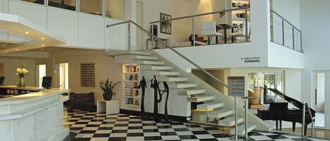 A spacious lobby with a checkered floor, a grand piano, and a staircase leading to a second floor with a library.