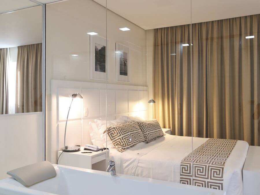 A modern hotel room with a large bed, a separate bathroom with a bathtub, a bedside table with a lamp, and a large mirror.