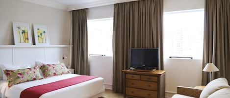 Una habitación de hotel con cama, televisor, cómoda, sofá y dos ventanas con cortinas.