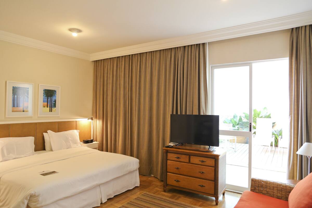 Hotelkamer met een bed, een televisie op een houten dressoir, een balkon met planten en een glazen schuifdeur.