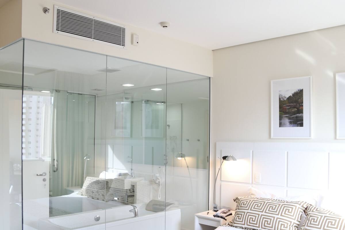 Een moderne slaapkamer met een douchecabine van glas, een wit bed met gestreepte kussens en een nachtkastje.