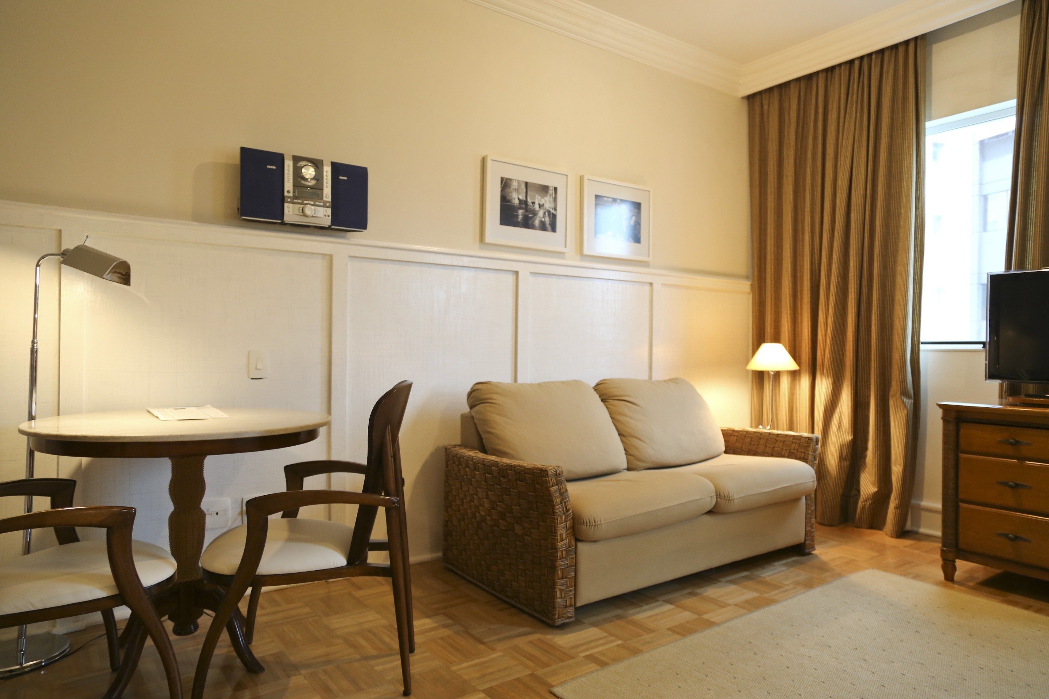 A hotel room with a round dining table, two chairs, a sofa, a TV stand, and a lamp.