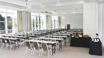 Uma sala de conferências com fileiras de mesas e cadeiras, grandes janelas e um palco.