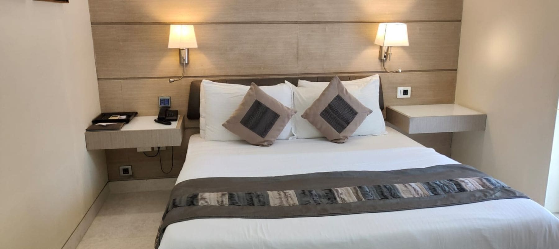 A hotel room with a bed, two bedside lamps, a nightstand with a phone, and a wall-mounted thermostat.