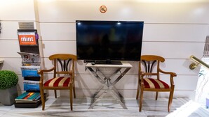 A room with a flat-screen TV mounted on the wall, two wooden chairs with red and beige striped cushions, a small table in front, and a magazine rack with various publications.