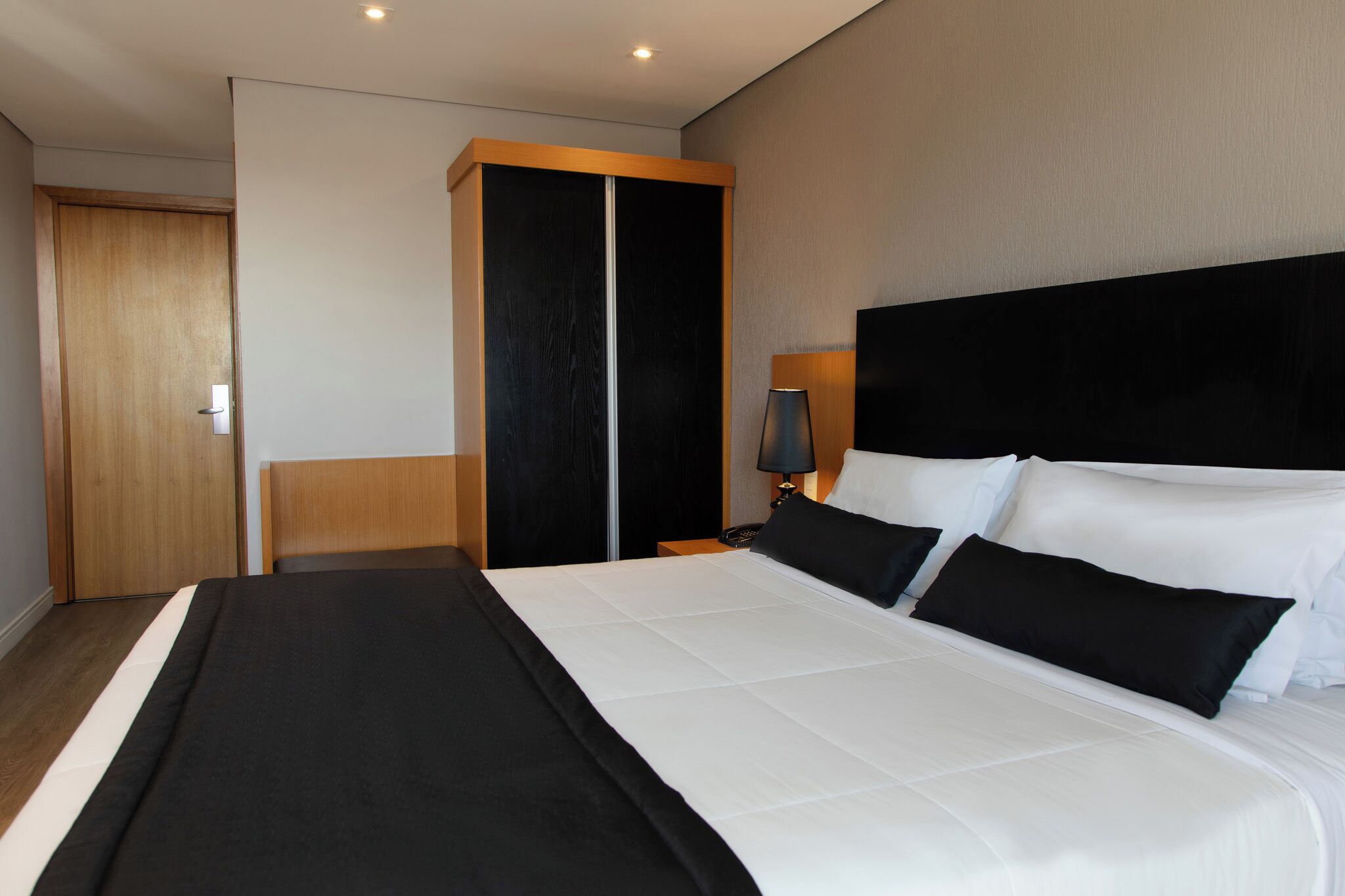 A modern hotel room with a large bed, a wardrobe, and a bedside table with a lamp.