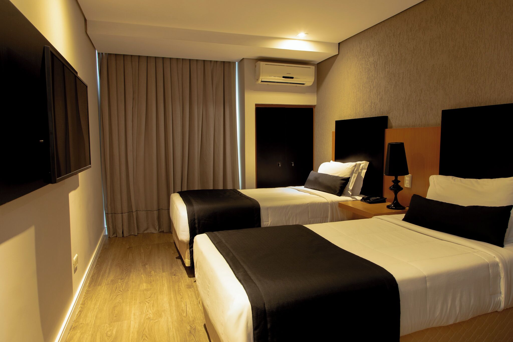 A hotel room with two beds, a TV, an air conditioning unit, and a nightstand with a lamp.