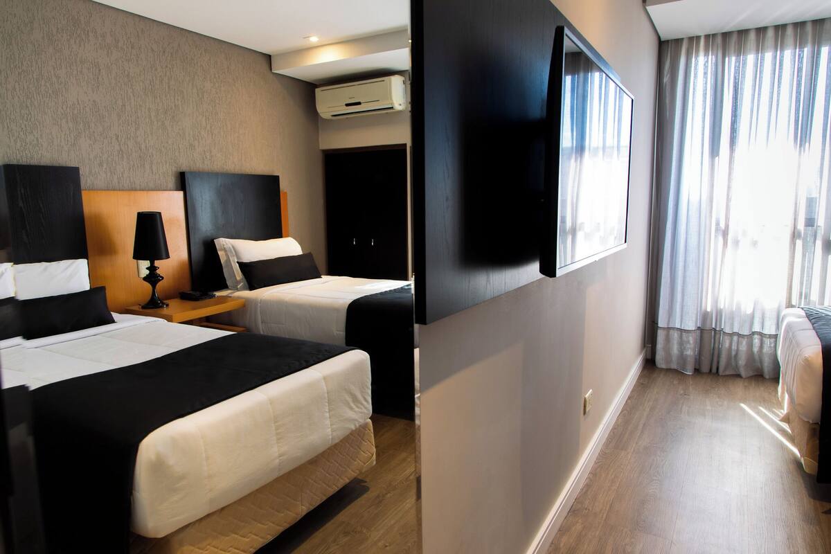 Habitación de hotel con dos camas, un televisor de pantalla plana, un equipo de aire acondicionado y un ventanal con cortinas.