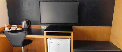 A compact room with a black desk, a black chair, a small white refrigerator, and a large flat-screen TV mounted on the wall.