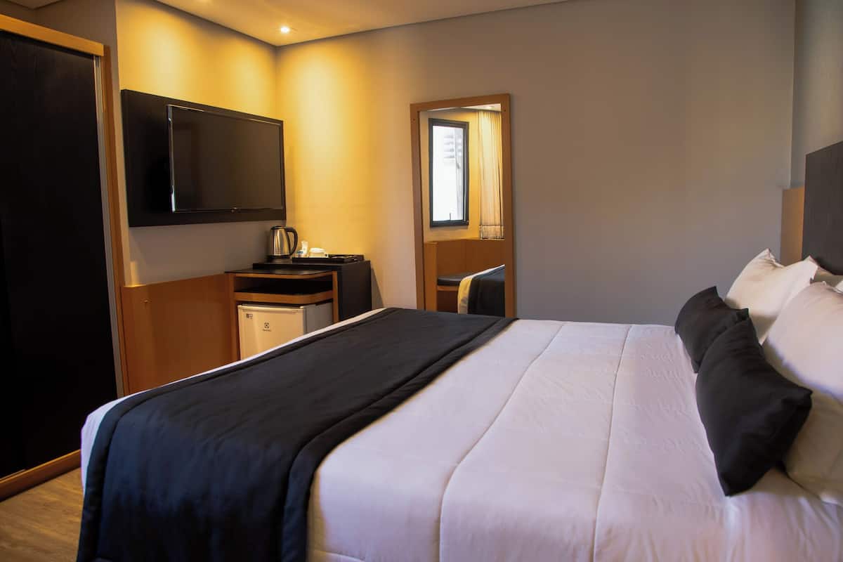 Habitación de hotel con una cama grande, un televisor de pantalla plana montado en la pared, un escritorio con un hervidor y una ventana con cortinas.
