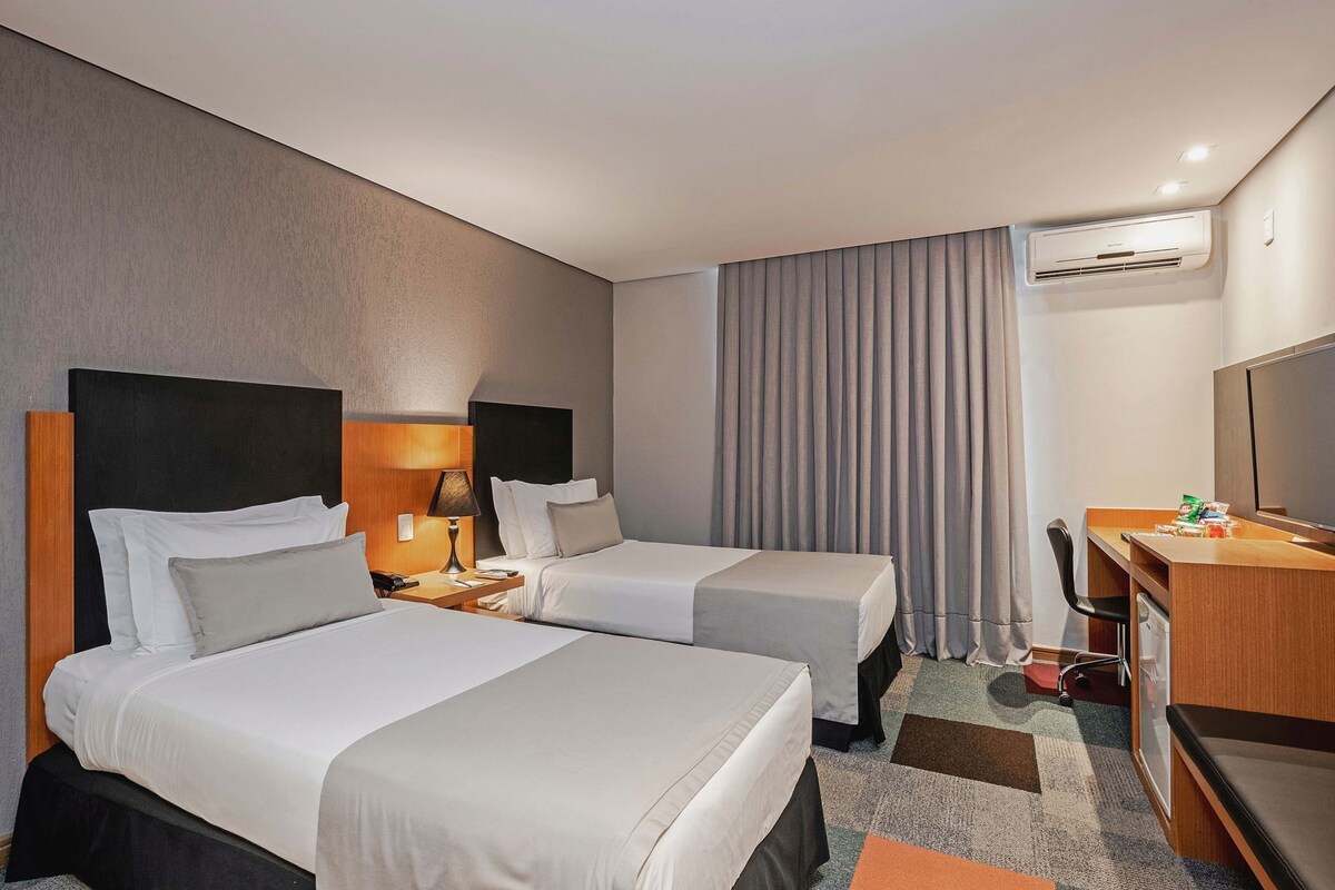 A hotel room with two beds, a desk, a chair, an air conditioning unit, and a television.