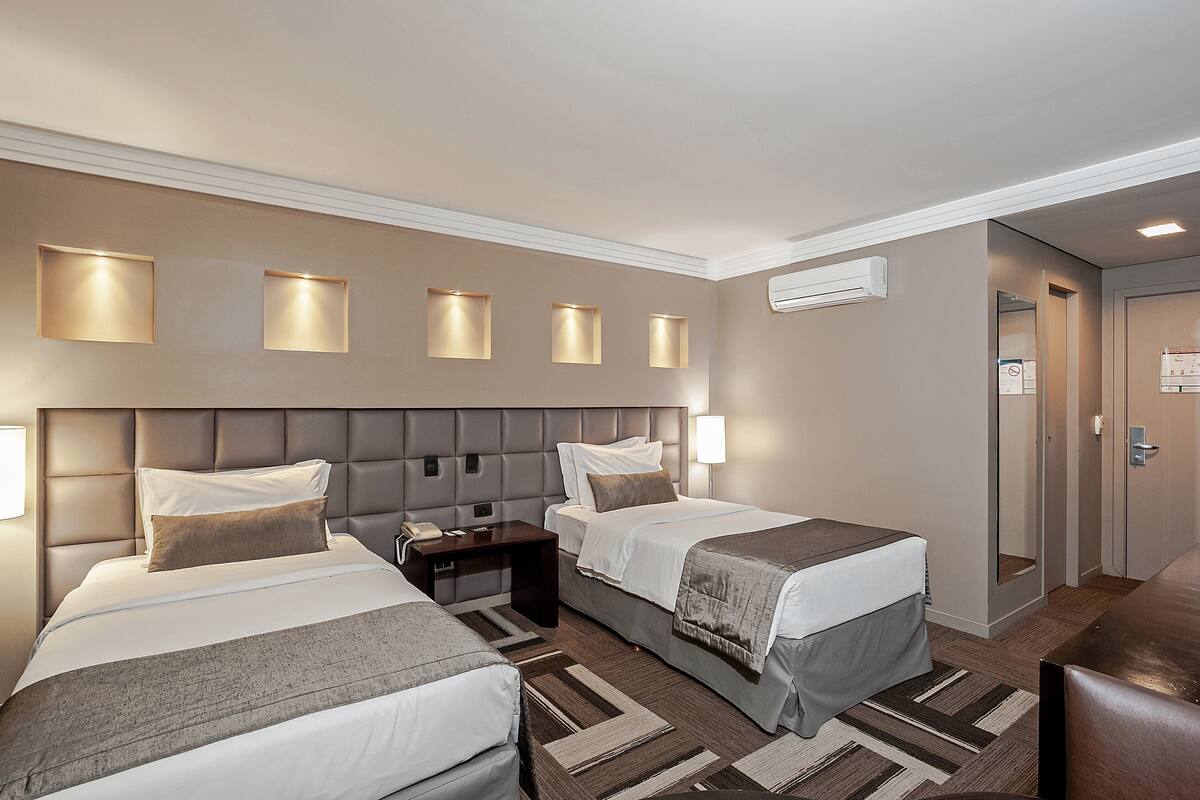 Habitación de hotel con dos camas, un escritorio y una mesita de noche. Hay luces fijadas a la pared sobre las camas y un aire acondicionado mural.
