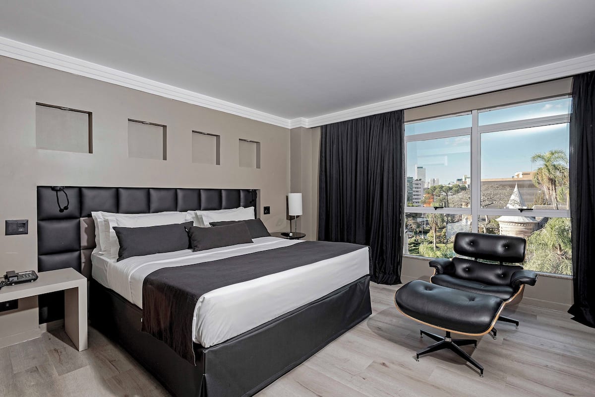 Habitación de hotel moderna con una cama grande, un escritorio, una silla y un amplio ventanal con vistas a la ciudad.