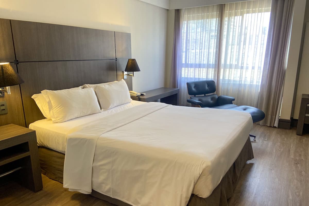 Quarto de hotel com uma cama grande, uma escrivaninha, uma cadeira e uma janela com cortinas.
