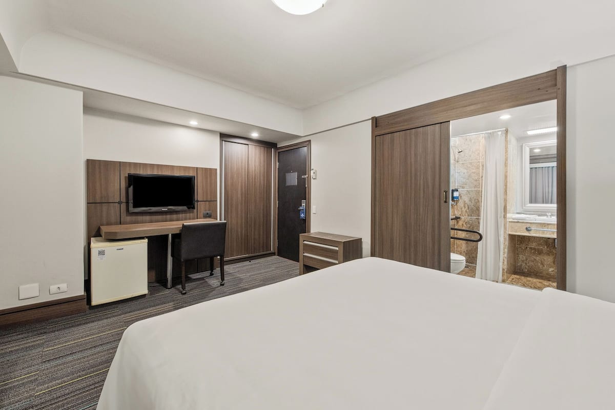 Quarto de hotel com cama, escrivaninha com cadeira, televisão, guarda-roupa e banheiro com chuveiro e vaso sanitário.