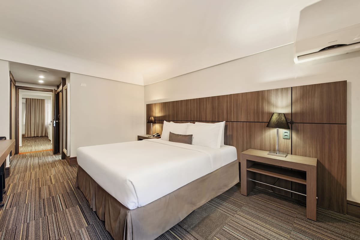 Quarto de hotel com uma cama grande, mesa de cabeceira com abajur, cabeceira de madeira e piso coberto por carpete.