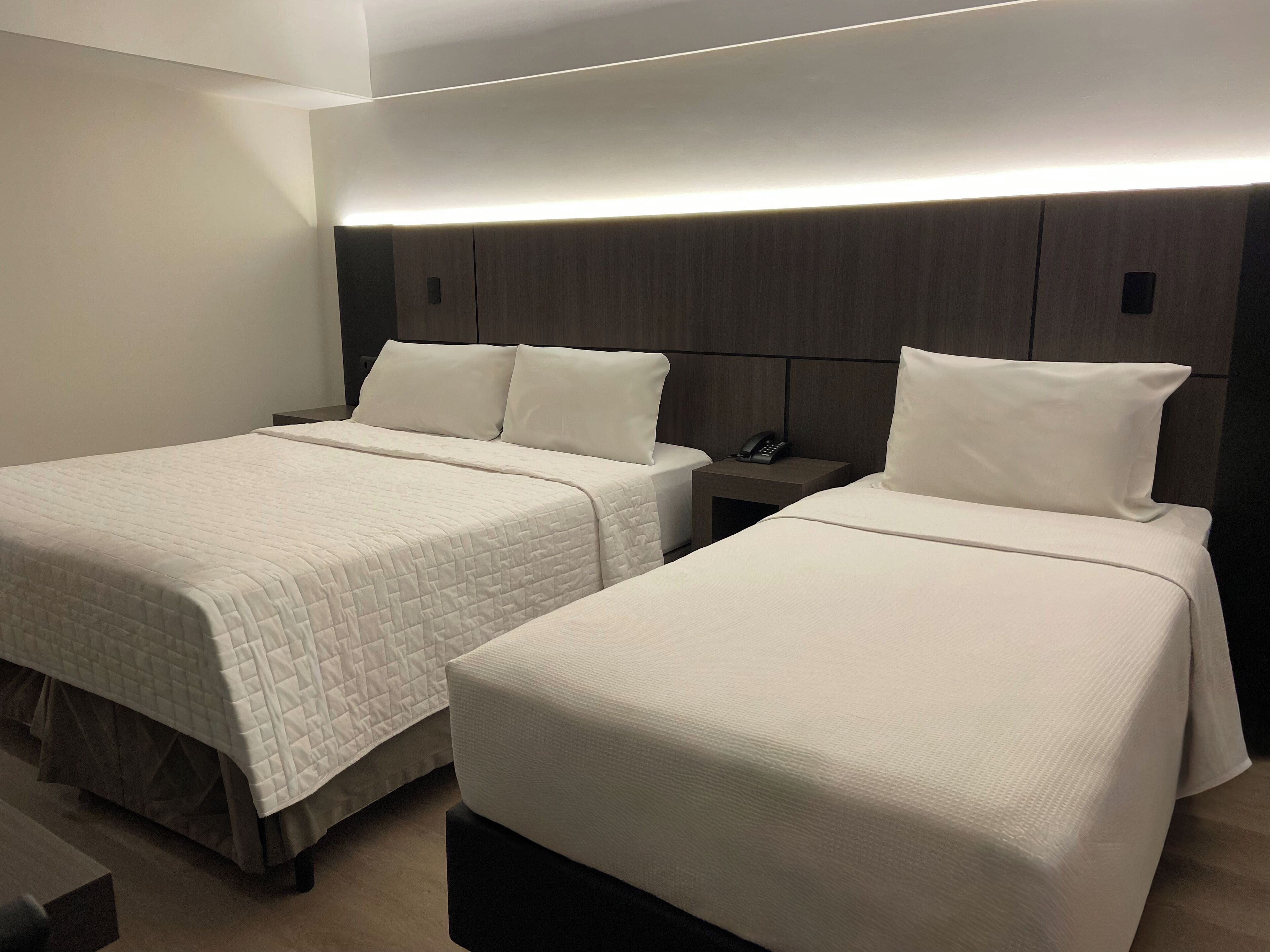 A hotel room with two beds, each with a white quilted bedspread and matching pillows. There is a bedside table with a phone on it, and the room has a modern design with a wooden headboard and a light strip above the beds.