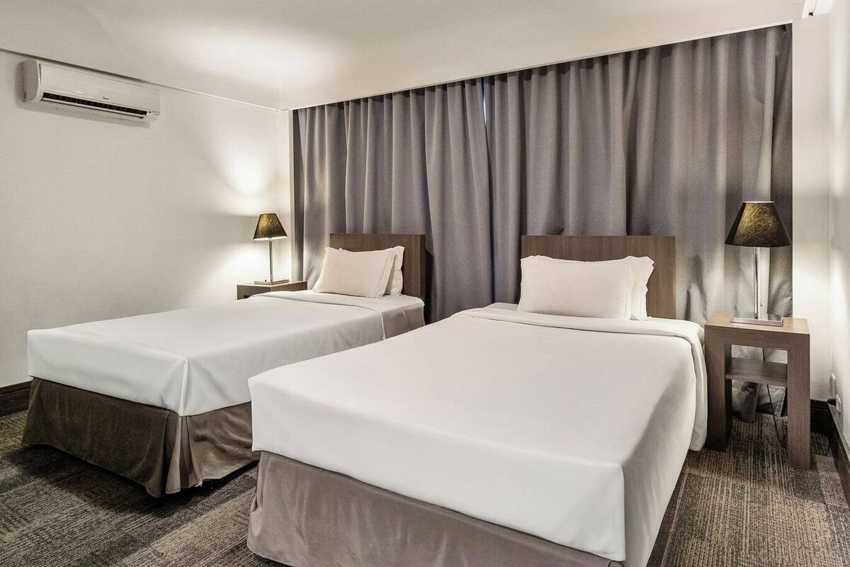 Quarto de hotel com duas camas, uma luminária no teto, ar-condicionado e uma janela com cortinas.