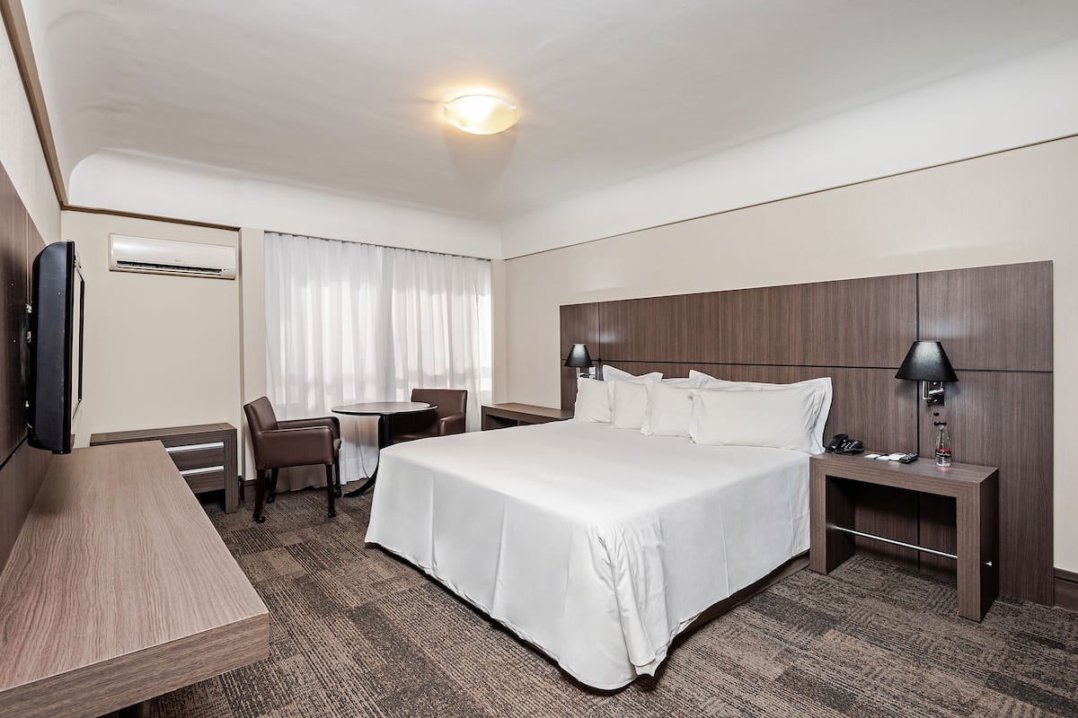 Quarto de hotel com uma cama grande, uma escrivaninha, uma cadeira, uma televisão, um abajur e um aparelho de ar condicionado.