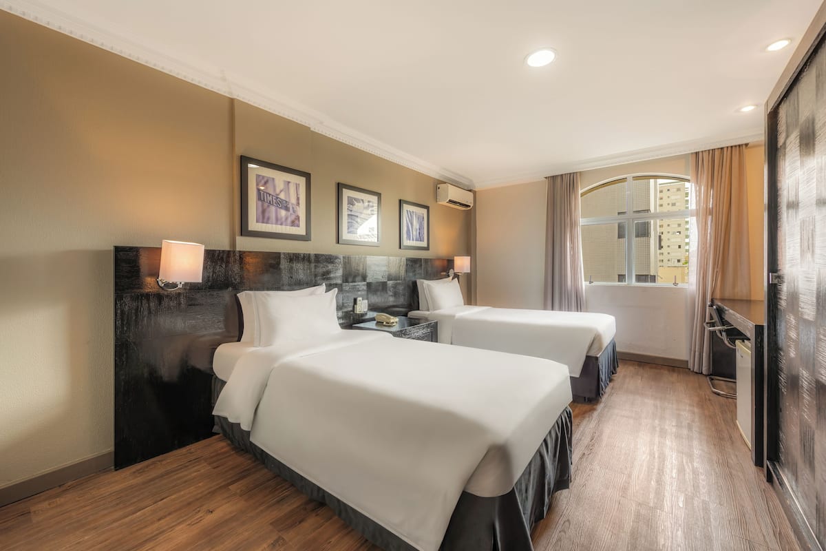 A hotel room with two beds, a wooden floor, a large window with curtains, and framed artwork on the wall.