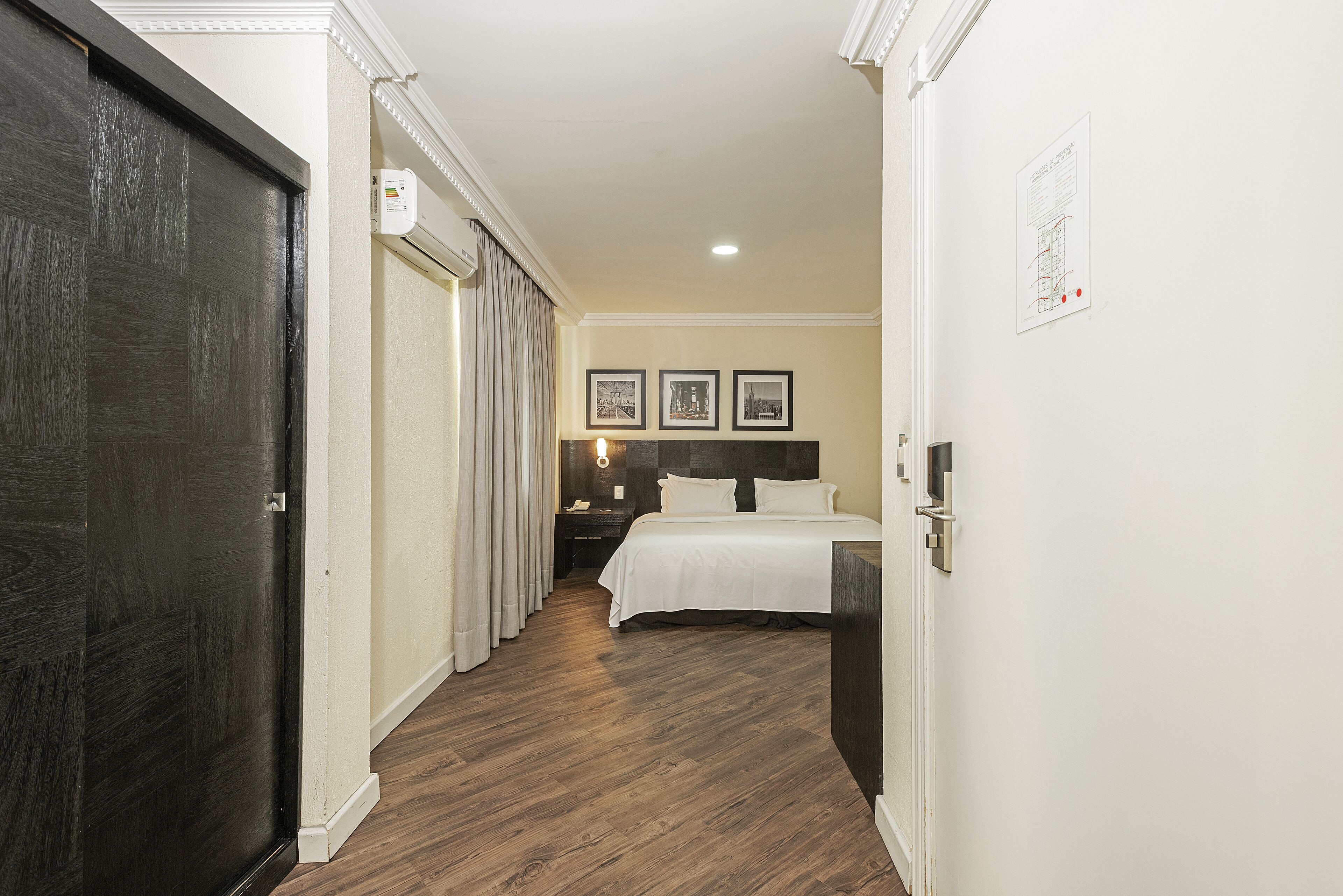 A hotel room with a wooden floor, a bed with white bedding, a nightstand, and a wall-mounted light fixture.