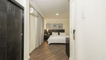 A hotel room with a wooden floor, a bed with white bedding, a nightstand, and a wall-mounted light fixture.