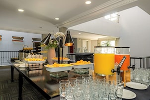 Disposição de buffet com diversos alimentos e um dispensador de suco, tudo cercado por copos e pratos.