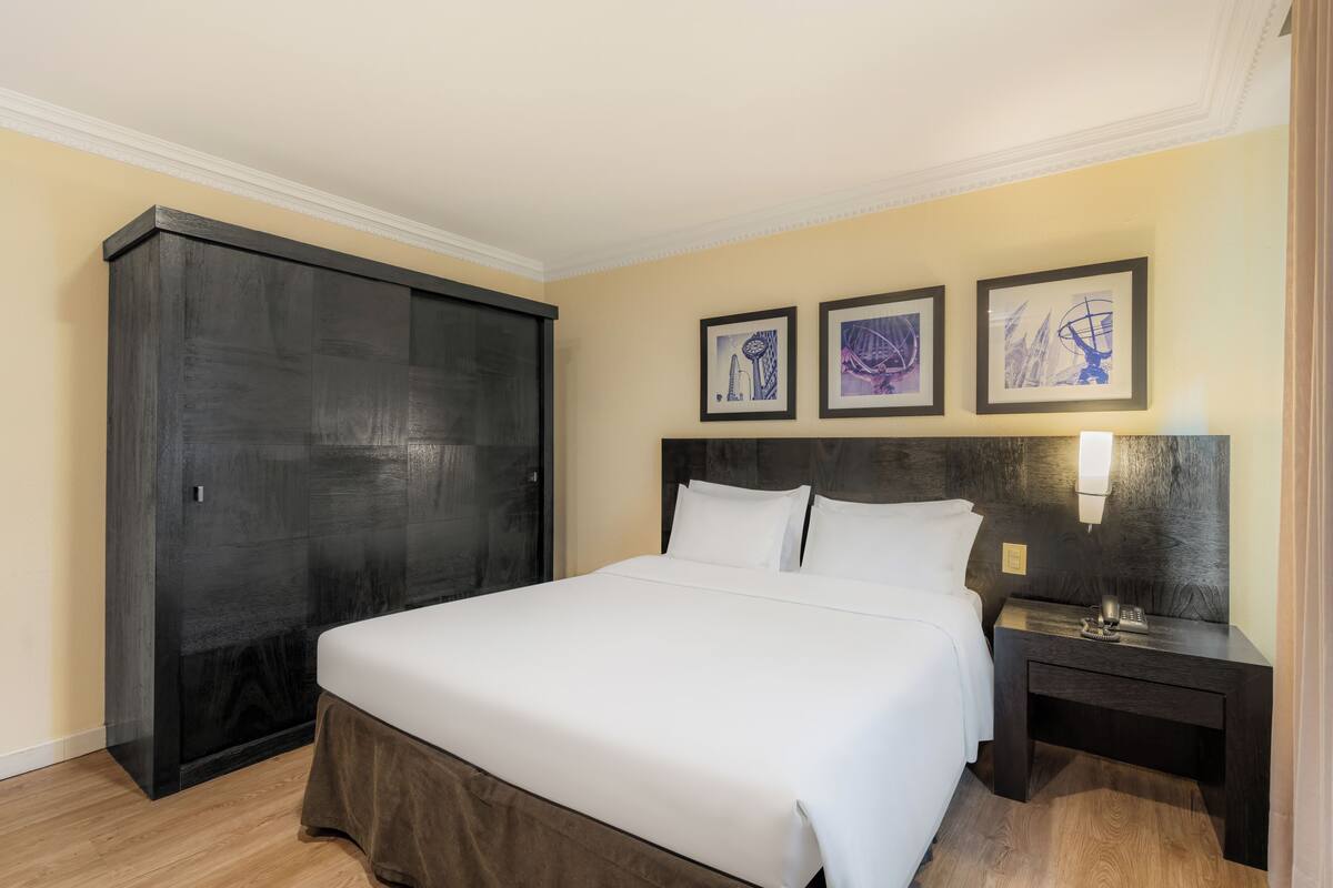 A hotel room with a large bed, a wardrobe, a nightstand, and three framed artworks on the wall.