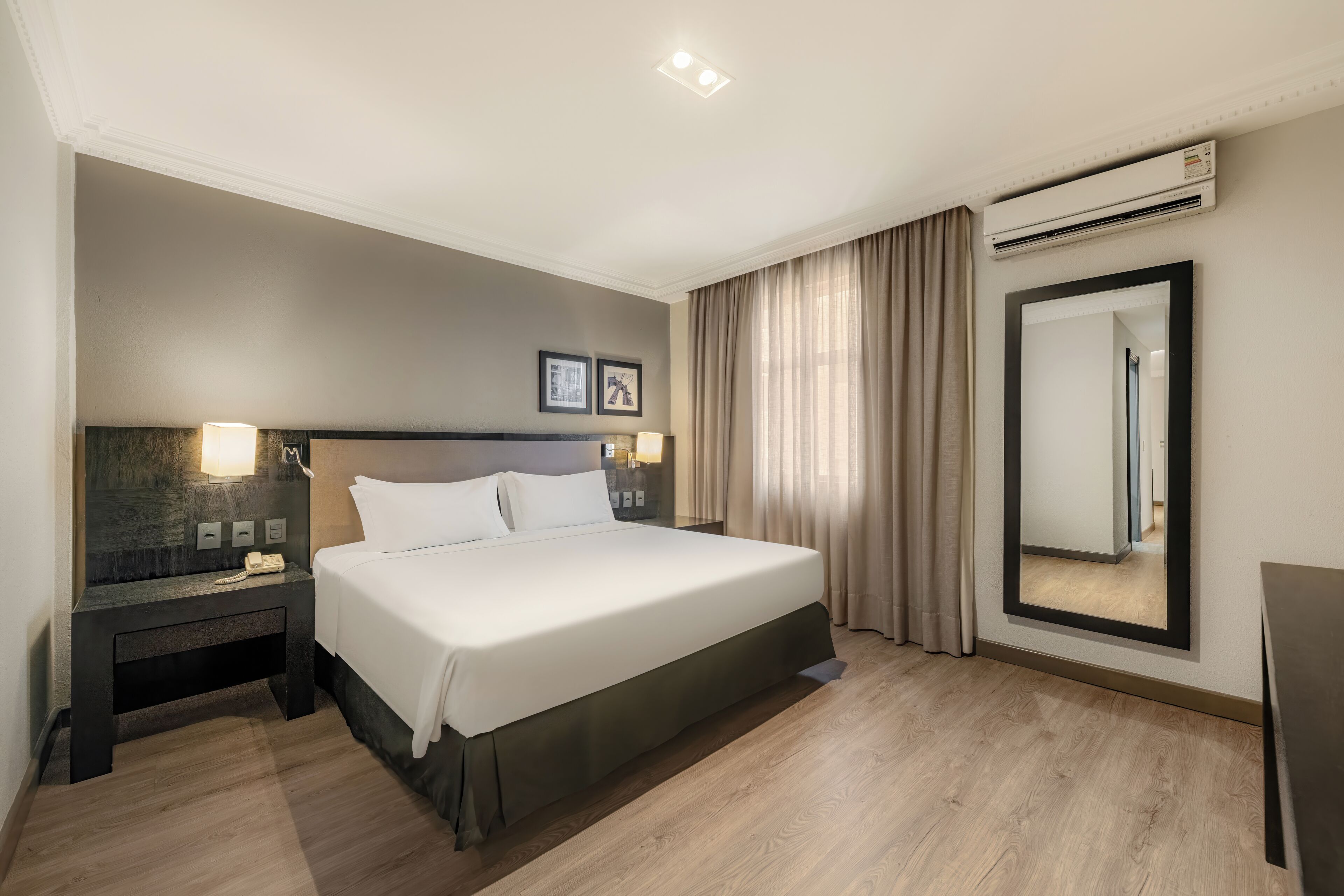 A modern hotel room with a large bed, bedside tables, a mirror, and an air conditioning unit.