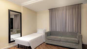 Habitación de hotel con una cama individual, un sofá gris, un espejo grande y una ventana con cortinas.