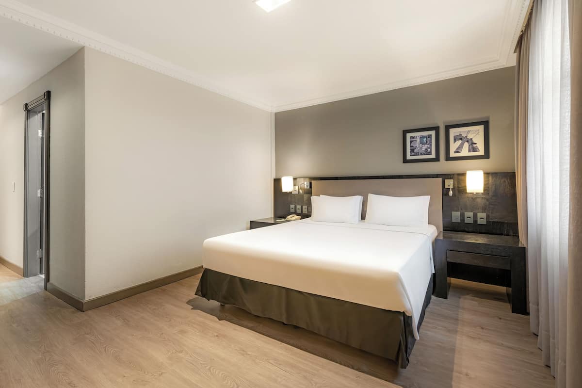 A hotel room with a large bed, two bedside tables, a nightstand, a bench, and a door.