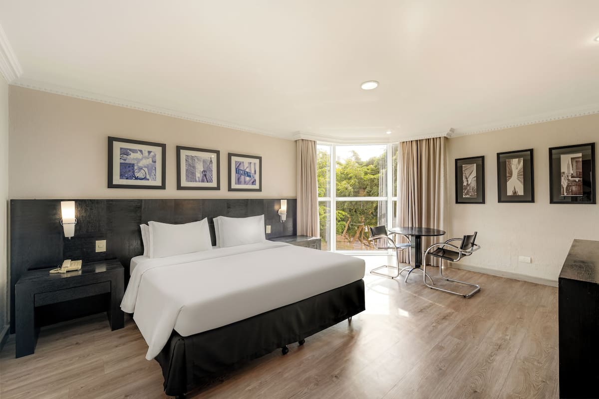 A modern hotel room with a large bed, a desk, and a chair. There are framed pictures on the wall and a window with curtains.