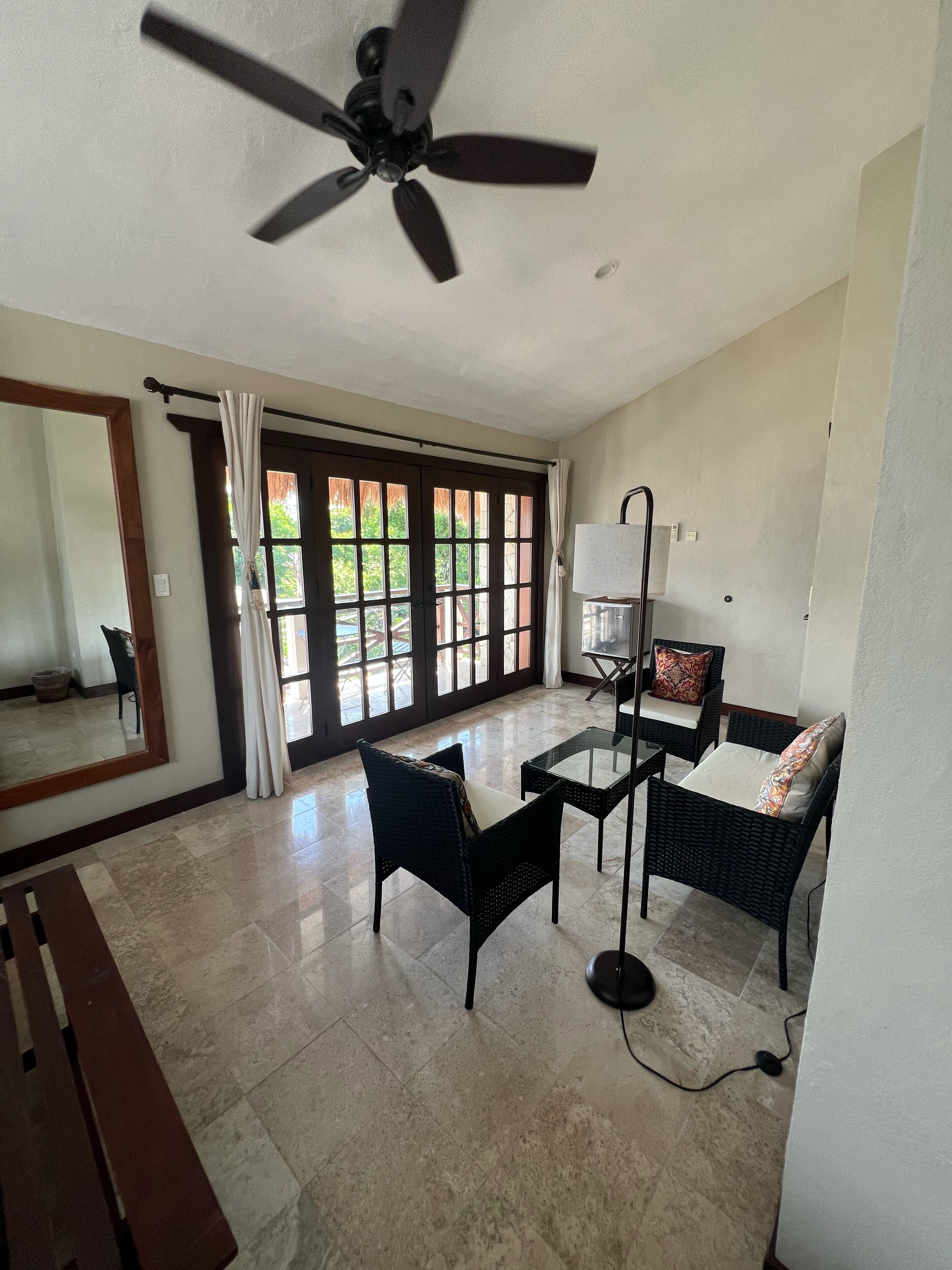 Una habitación amplia con ventilador de techo, ventanas grandes con cortinas, una zona de estar con muebles de mimbre y piso de baldosas.