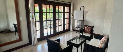 Una habitación amplia con ventilador de techo, ventanas grandes con cortinas, una zona de estar con muebles de mimbre y piso de baldosas.