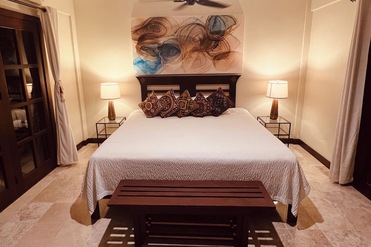 Une chambre d’hôtel avec un grand lit, deux lampes de chevet, un ventilateur de plafond et un tableau au-dessus du lit.