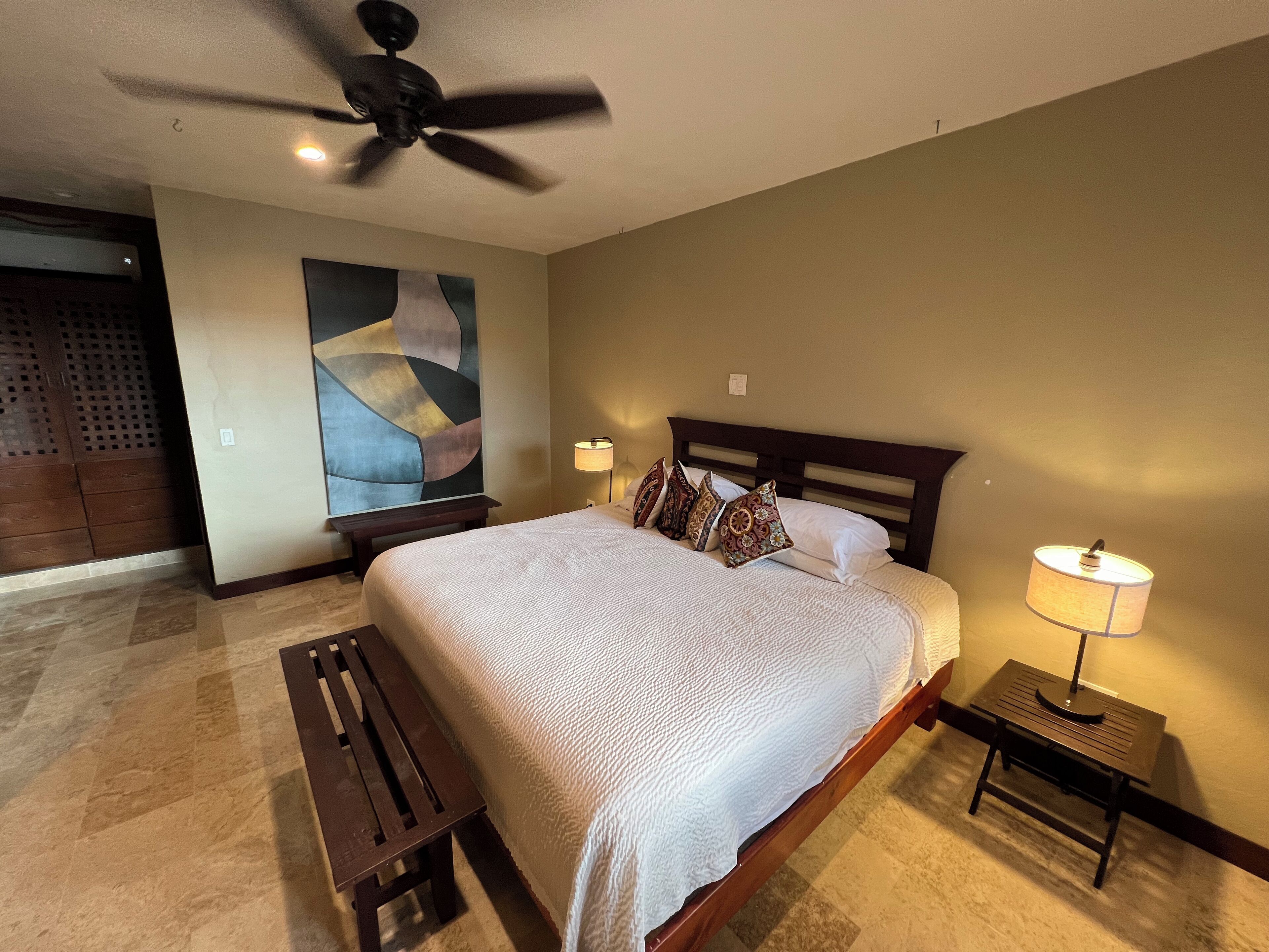 A bedroom with a large bed, a ceiling fan, a nightstand with a lamp, and a bench.
