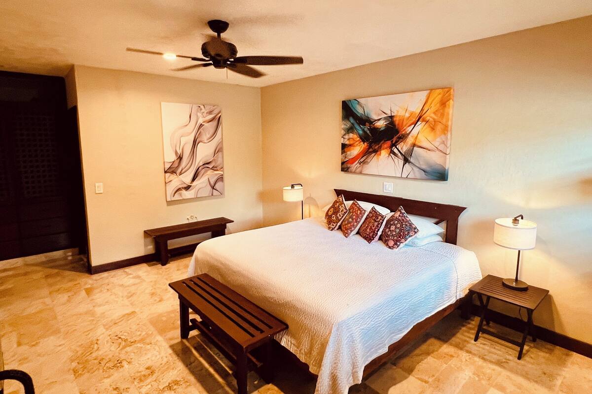 Une chambre à coucher comprenant un lit, un ventilateur de plafond, deux tables de chevet et des œuvres d’art accrochées aux murs.