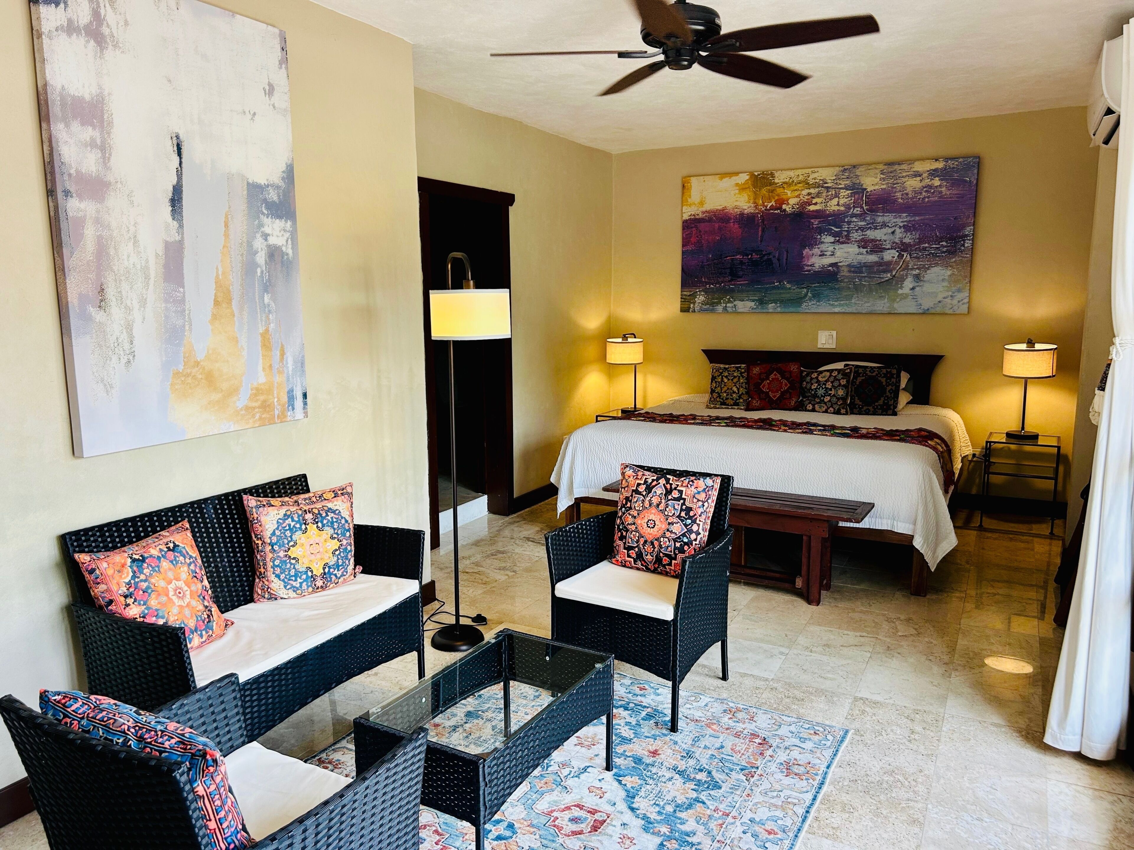Une chambre d’hôtel avec un lit, deux chaises, une table, un ventilateur de plafond, un tableau et une lampe.